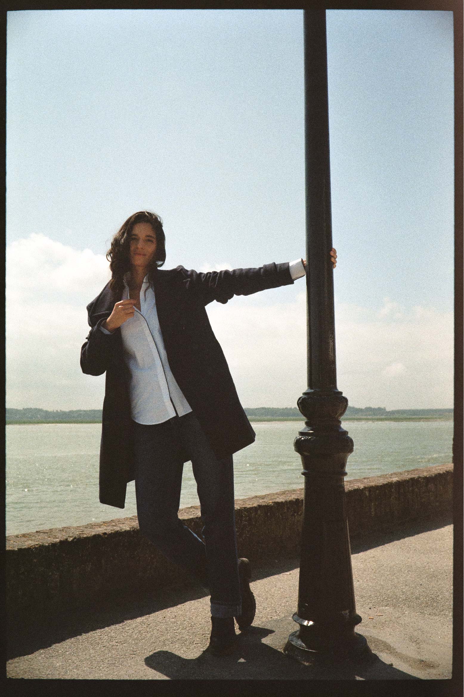 Photo prise de plain pied, mannequin s’appuie sur un lampadaire, elle porte une chemise en popeline blanche et avec un passepoil bleu marine le long de la gorge de boutonnage ainsi que sur les poignets.