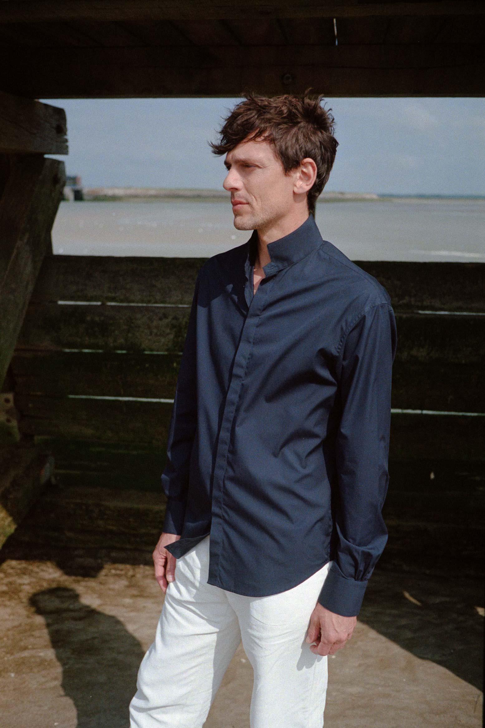 BOURRIENNE PARIS X: BOURRIENNE PARIS X: Photo prise de face, le mannequin regarde à l’horizon et porte une chemise en popeline bleu marine. La chemise tombe sur ses hanches, le col est haut, les poignets sont classiques et ont des fronces.