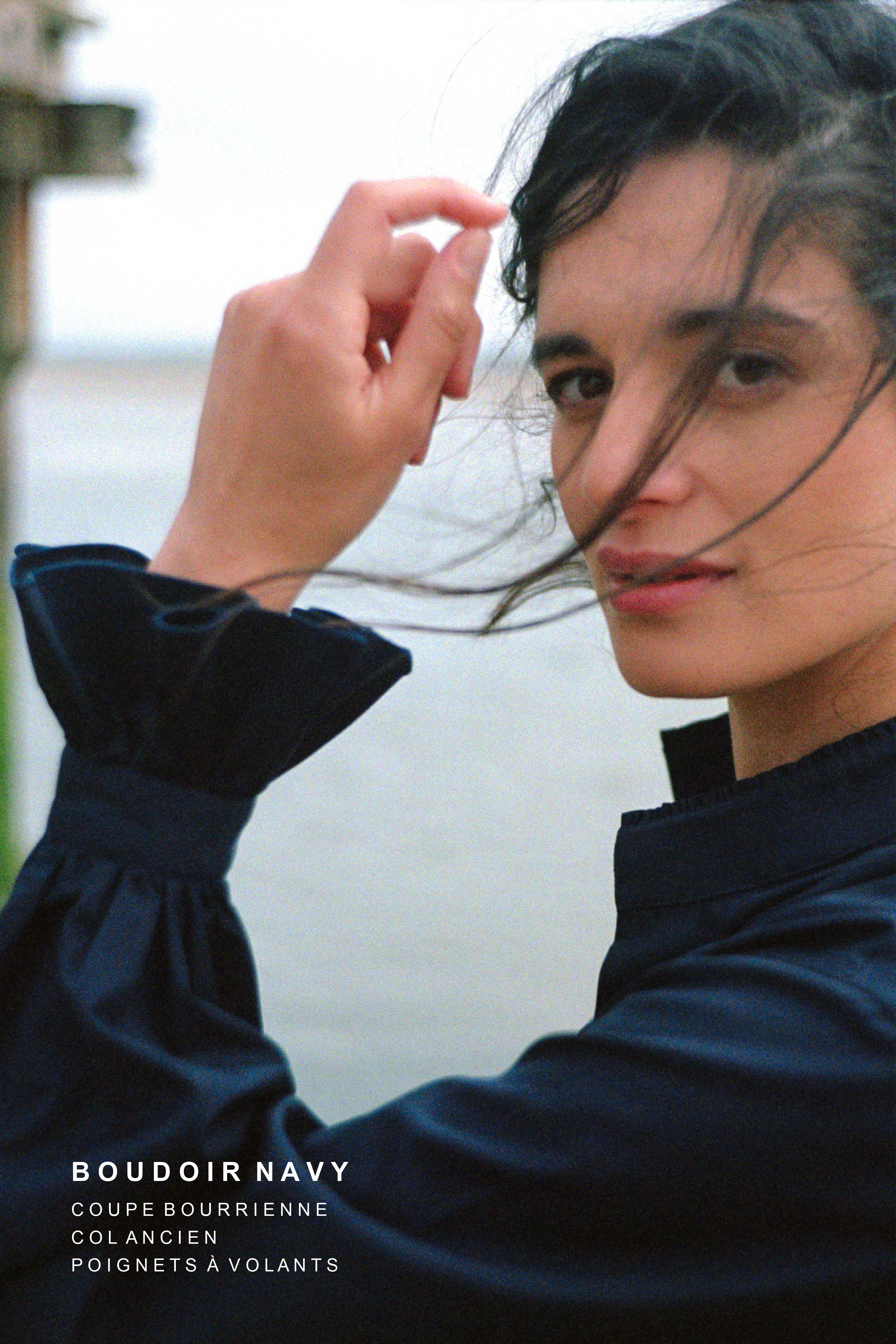 BOURRIENNE PARIS X: Photo de profil, le mannequin à des mèches de cheveux sur son visage et montre son avant bras de manière à ce qu’on puisse apercevoir les détails des manches bouffantes et des volants brodés au niveau des poignets.