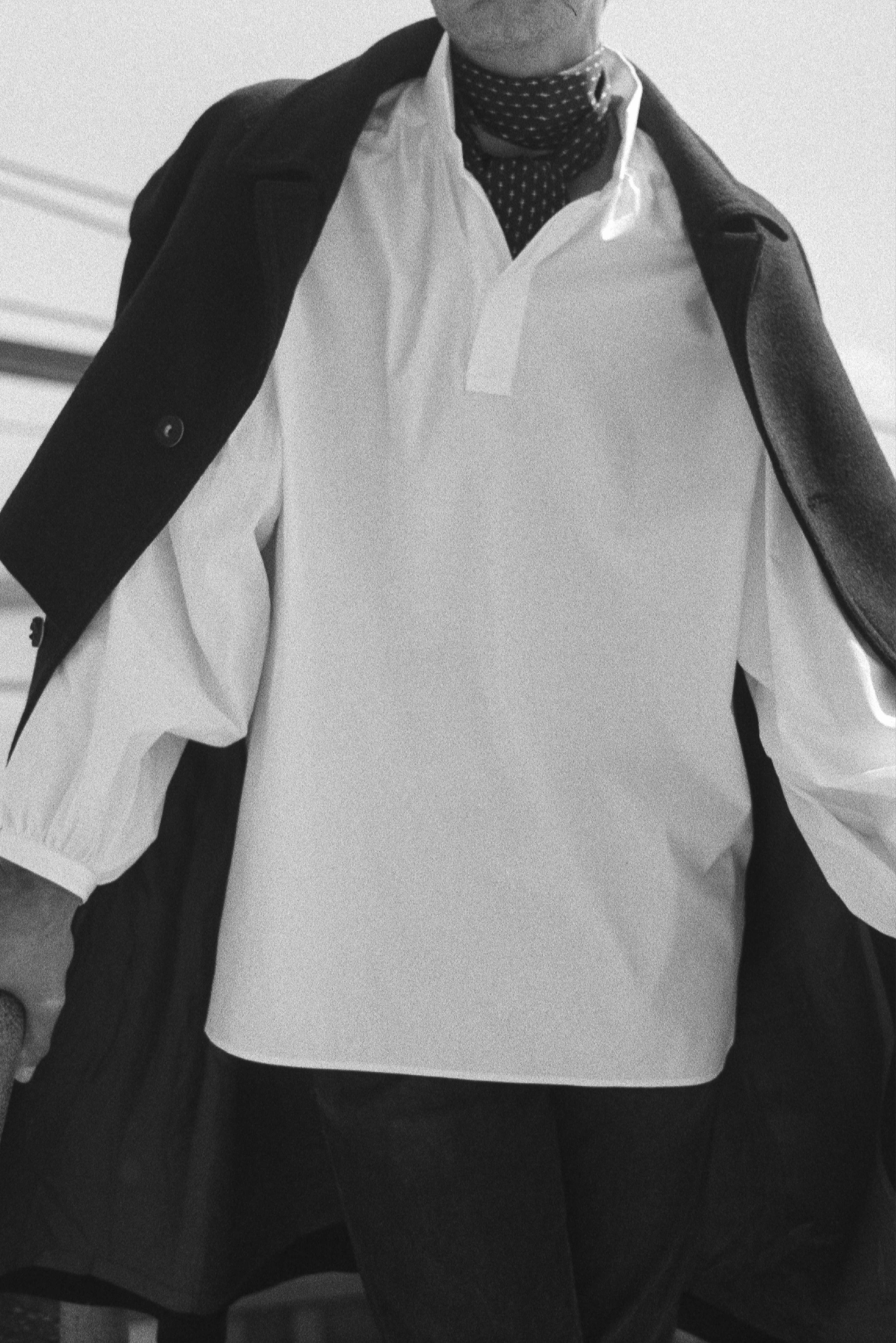 BOURRIENNE PARIS X: Photo prise de face, le mannequin porte une chemise en popeline blanche et un long manteau bleu marine. La chemise a de grandes manches napoléoniennes, un col haut et des poignets fins et resserrés. La photo est en noir et blanc.