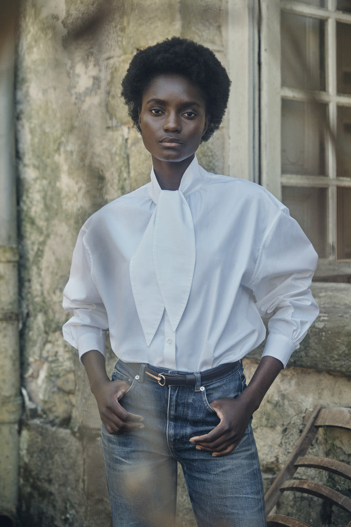 Femme portant la chemise MERVEILLEUSE, rentrée dans un jean bleu, posant devant une maison en pierre.