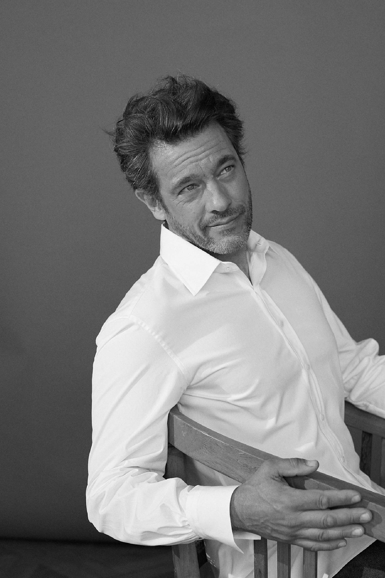 Photo noir et blanc plan poitrine d'un homme portant une chemise en popeline blanche double retors, col français et poignets bourrienne. L'homme est assis, ses épaules sont légèrement vers l’arrière et son bras droit est collé au dossier de sa chaise on voit les details de sont poignet droit avec les fronces qui débute sur la chemise du poignet.