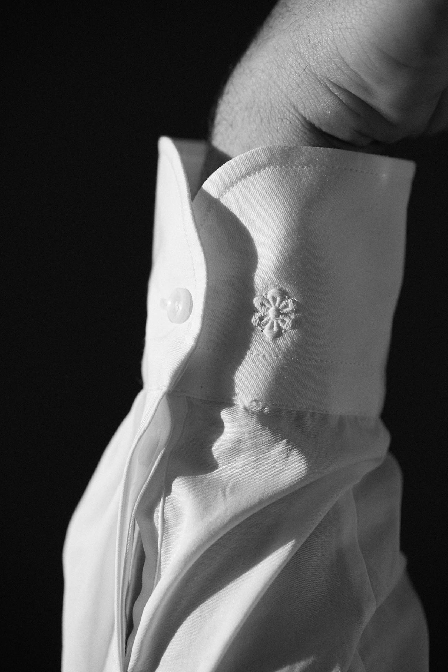 Photo gros plan en noir et blanc d'un poignet d'une chemise blanche classique en popeline double retors. En deuxième plan le logo brodé de la fleur Bourrienne.