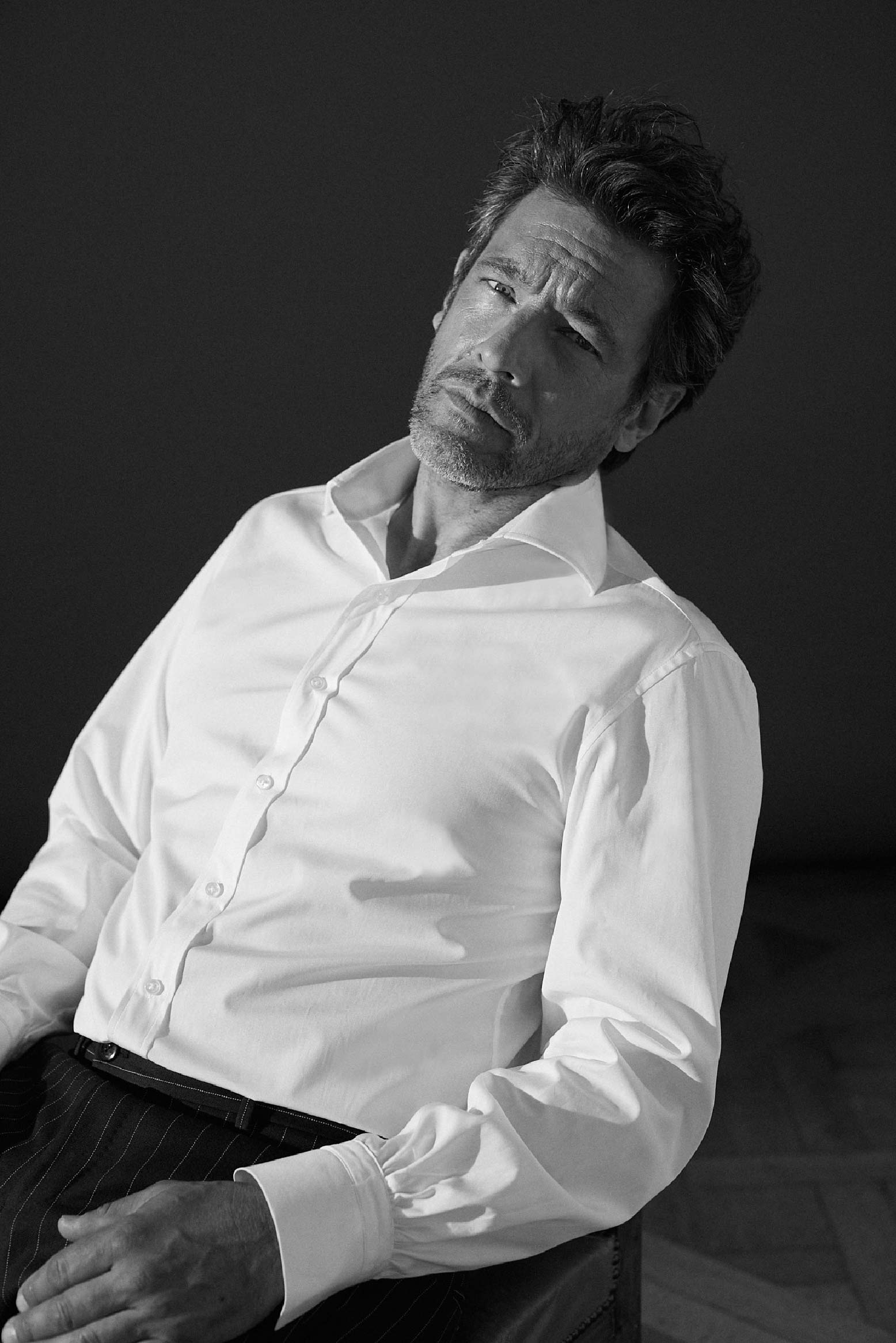 Photo plan américain en noir et blanc d'un homme assis sur une chaise il porte une chemise en popeline blanche italienne double retors, col italien et poignet Bourrienne. Son dos penche vers l’arrière, son visage fixe l'objectif, le premier bouton de sa chemise est ouvert. Ses mains sont posées sur ses genoux , on voit en premier plan le poignet Bourrienne de sa chemise.