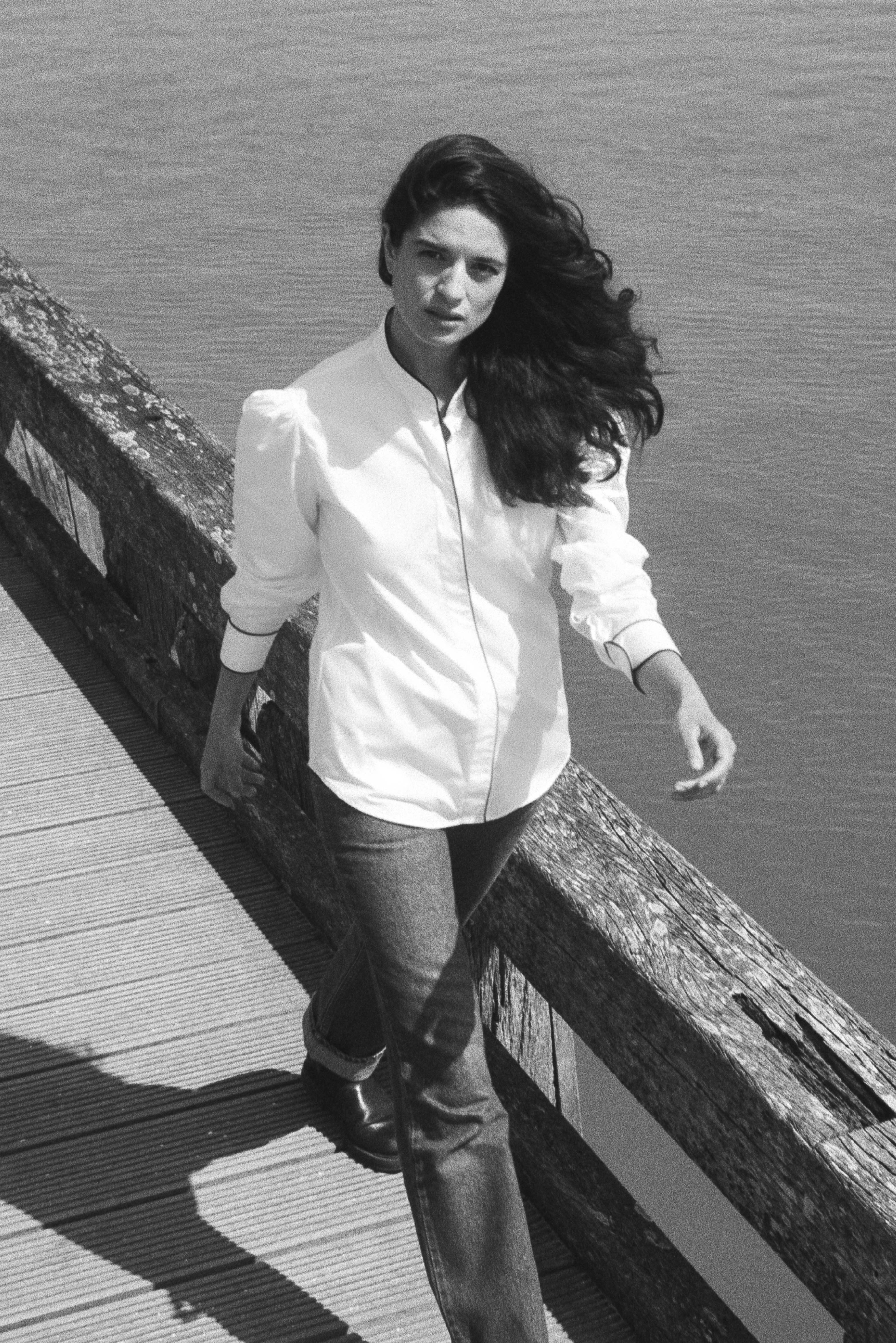 Photo prise de haut, le mannequin marche et regarde la caméra, la phot est en noir et blanc, le mannequin porte une chemise en popeline blanche, on aperçoit le détail des épaules bouffantes grâce à leurs fronces.