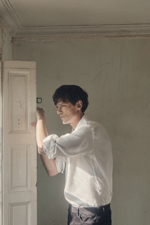 Homme de profil, les bras posés sur les volets d’une fenêtre ancienne, vêtu d’une chemise PALETTE en soie blanche à coupe Bourrienne et col français ouvert. Les manches sont subtilement retroussées, mettant en valeur la fluidité satinée du tissu et la finesse des poignets classiques. La lumière naturelle qui filtre à travers les carreaux illumine la texture délicate de la soie, contrastant avec l’enduit écaillé du mur.