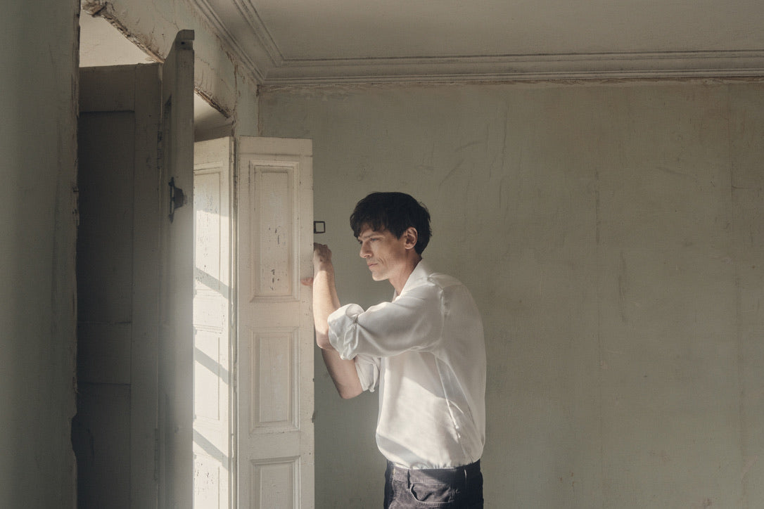 Homme de profil, les bras posés sur les volets d’une fenêtre ancienne, vêtu d’une chemise PALETTE en soie blanche à coupe Bourrienne et col français ouvert. Les manches sont subtilement retroussées, mettant en valeur la fluidité satinée du tissu et la finesse des poignets classiques. La lumière naturelle qui filtre à travers les carreaux illumine la texture délicate de la soie, contrastant avec l’enduit écaillé du mur.