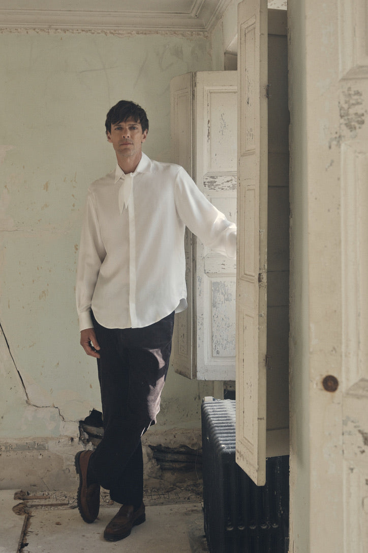 Homme debout, la main droite posée sur un volet ancien, vêtu de la chemise PALETTE en soie ivoire à coupe Bourrienne et col français noué en un large nœud décontracté, manches légèrement retroussées révélant les poignets classiques. Le tissu satiné épouse la silhouette et capte la lumière filtrée par la fenêtre à petits carreaux, créant un délicat jeu d’ombres et de reflets. En contraste, un pantalon en velours côtelé sombre et le mur patiné aux crépis écaillés soulignent l’élégance fluide de la tenue.