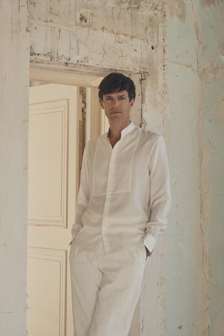 Homme debout dans l’embrasure d’une porte patinée, mains en partie dans les poches d’un pantalon clair, portant la chemise NOCTURNE en soie blanche à coupe ajustée et col officier ouvert. Le plastron revisité en nid d’abeille se détache sur la poitrine et les poignets Bourrienne signature, subtilement mis en valeur par les manches légèrement retroussées. Le mur usé au crépi patiné et la lumière tamisée mettent en relief la fluidité et l’éclat délicat de la soie.