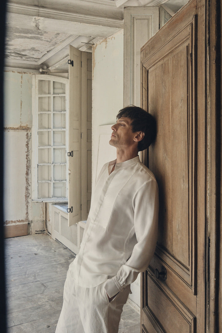 Homme de profil trois-quarts, adossé à une porte en bois patiné et mains partiellement dans les poches d’un pantalon blanc cassé, portant la chemise NOCTURNE en soie ivoire à coupe ajustée et col officier ouvert. Le plastron avant en nid d’abeille se déploie sur la poitrine tandis que les poignets Bourrienne, finement froncés et fermés par un bouton nacré, ressortent sous les manches longues. La lumière douce du jour, filtrée par une fenêtre à petits carreaux, fait ressortir l’éclat satiné de la soie.