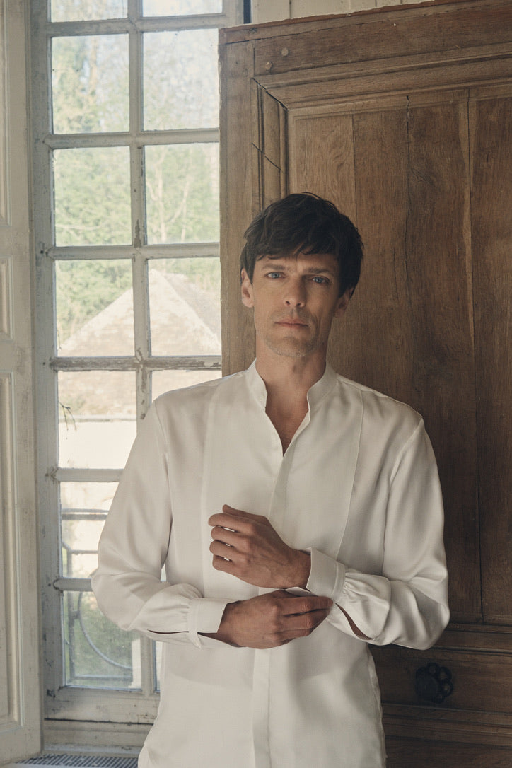 Homme debout dans un embrasure de fenêtre à petits carreaux, appuyé contre un vieux panneau de bois, ajustant délicatement le poignet de sa chemise NOCTURNE en soie blanche. Le col officier s’ouvre en V sur un plastron revisité en nid d’abeille, tandis que les poignets Bourrienne signature, finement froncés, soulignent l’élégance du tissu fluide. La lumière douce du jour révèle la texture satinée de la soie et le contraste entre le décor patiné et la coupe ajustée.