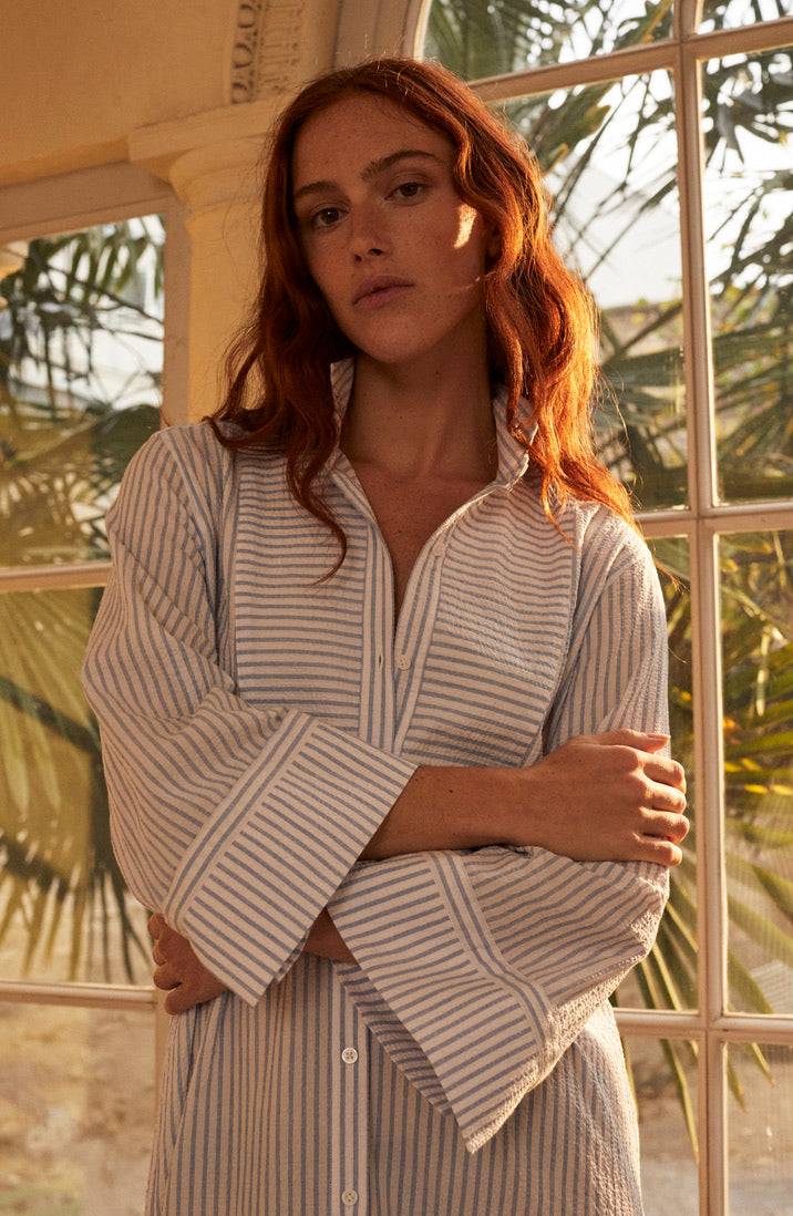 Photo plan américain d'une femme femme rouse debout, les bras croisés. Elle porte une robe chemise ROSEE, qui est coupée dans un tissu seersucker rayé bleu et blanc de haute qualité.
Elle s'inspire des détails emblématique de la Maison Bourrienne Paris X: un col officier haut, plastron et poignets reprenant les iconiques de la Maison. Ses fentes sur le côté accentuent une silhouette féminine.