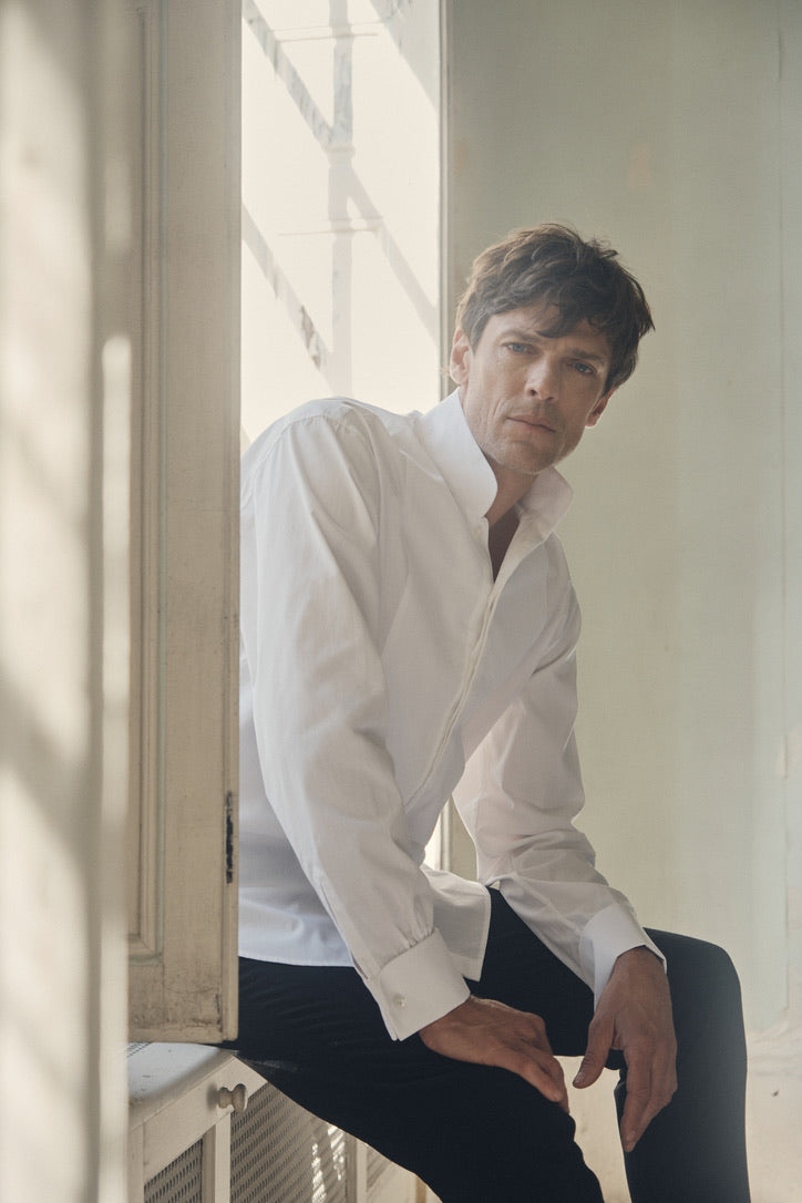 Photo plan américain d'un homme qui est assit, le visage face à la caméra, sa main gauche rentrée dans son pantalon, l'autre posé sur une fenêtre. Il porte une chemise blanche, coupe Bourrienne, poignets Français, col haut officier avec un plastron.
