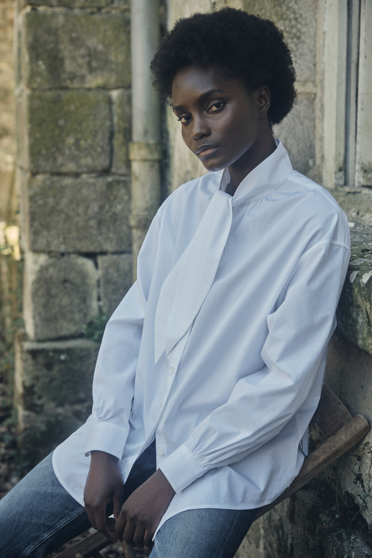 Femme portant la chemise blanche MERVEILLEUSE, avec col foulard posant devant une maison en pierre, plan américain.