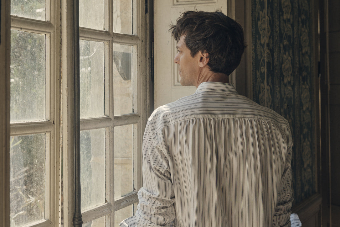 Homme de dos, debout près d’une fenêtre à petits carreaux, vêtu de la chemise ÉPICURIEN Rayée Kaki ; on distingue la coupe Bourrienne ample et les fronces dans le dos, sublimées par le col officier biseauté, dans une lumière douce filtrant l’intérieur ancien.