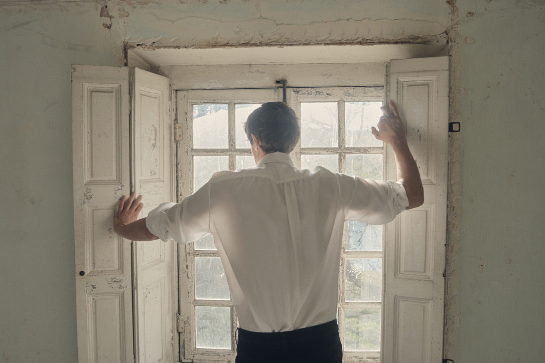 Homme de dos, bras écartés pour ouvrir les volets d’une fenêtre ancienne, vêtu de la chemise PALETTE en soie blanche à coupe Bourrienne et col français. Le dos dévoile un empiècement plissé subtilement froncé, rehaussé par la fluidité satinée du tissu. La lumière du jour pénètre à travers les carreaux, soulignant le contraste entre l’élégance du vêtement et le charme patiné du décor.