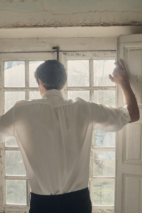 Homme de dos, bras écartés pour ouvrir les volets d’une fenêtre ancienne, vêtu de la chemise PALETTE en soie blanche à coupe Bourrienne et col français. Le dos dévoile un empiècement plissé subtilement froncé, rehaussé par la fluidité satinée du tissu. La lumière du jour pénètre à travers les carreaux, soulignant le contraste entre l’élégance du vêtement et le charme patiné du décor.