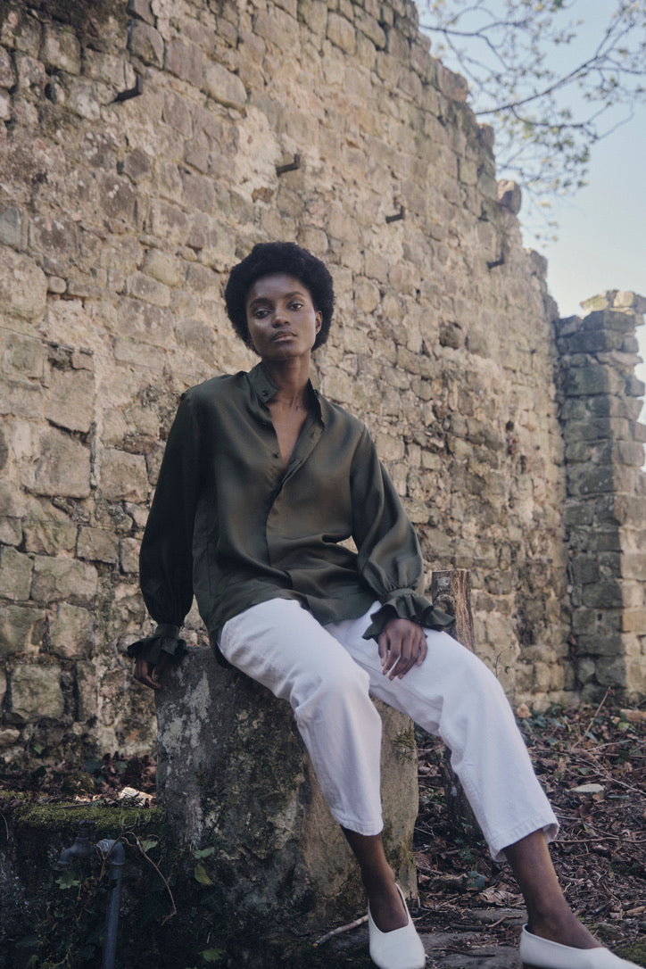 Chemise BOUDOIR Soie Kaki portée par une mannequin assise sur une pierre dans un jardin, vue en contre-plongée.