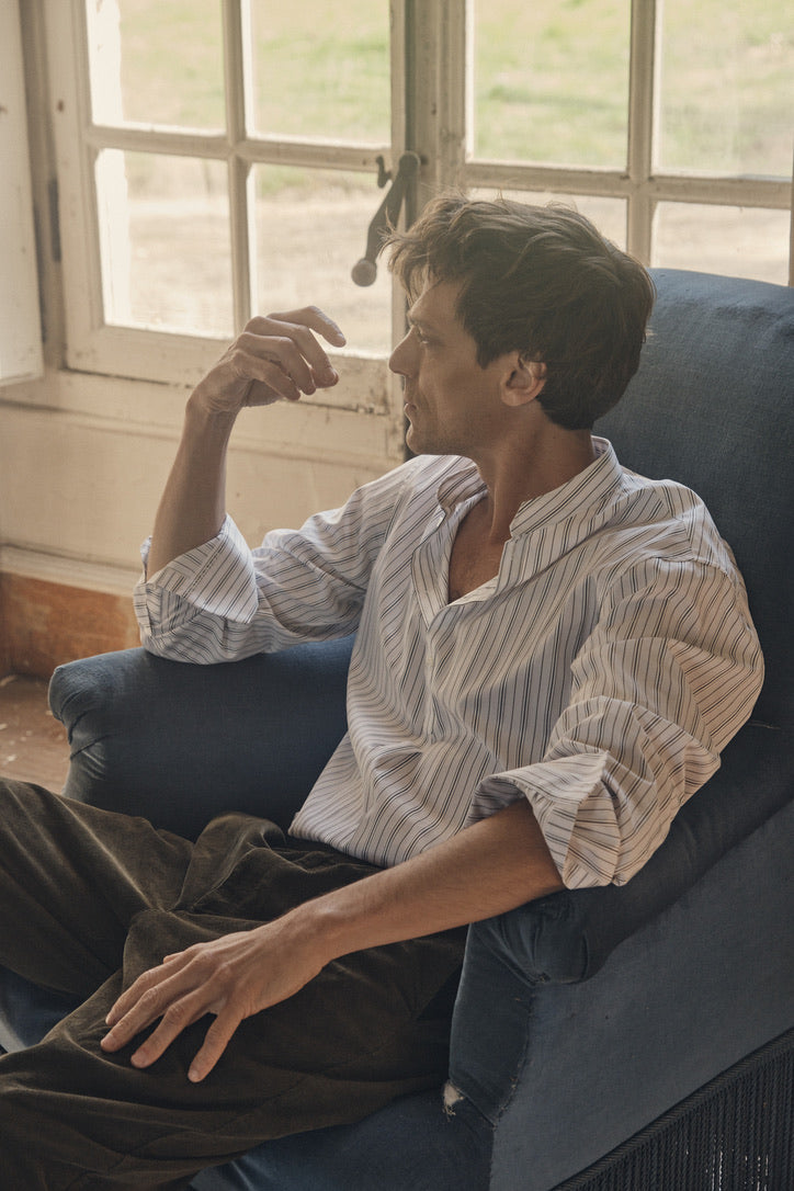 Homme de profil assis dans un fauteuil bleu face à une fenêtre à petits carreaux, portant la chemise ÉPICURIEN Rayée Kaki en popeline de coton : coupe Bourrienne ample, col officier biseauté et poignets classiques retroussés, avec un pantalon en velours côtelé kaki.