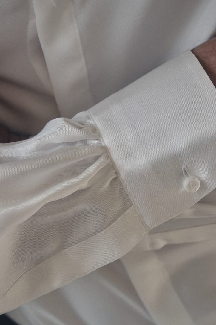 Gros plan sur le poignet gauche enchevêtré dans la manche retroussée de la chemise PALETTE en soie blanche, révélant un poignet classique fermé par un bouton nacré. Le tissu satiné, fluide et légèrement plissé sous la couture, capte la lumière naturelle pour faire ressortir son éclat délicat. La couture invisible du plastron et la finesse des fronces soulignent la qualité artisanale de la pièce.