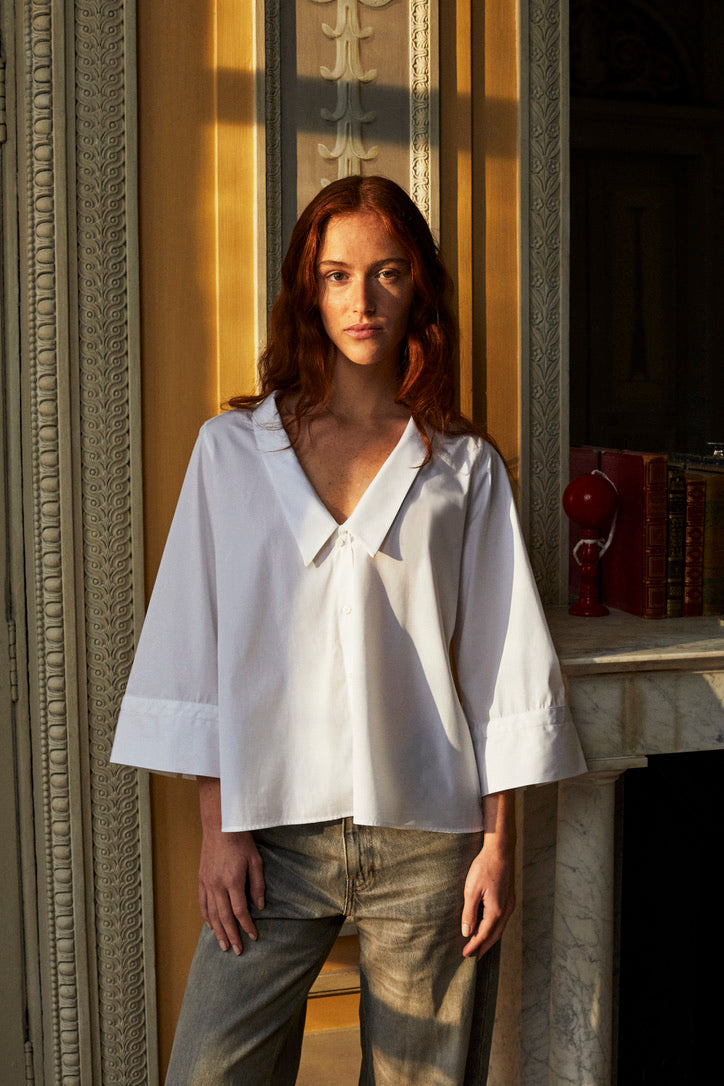 Photo plan américain d'une femme rousse les cheveux détachés, elle est debout derriere un mur et se trouve à côté d'une cheminée. Elle porte un jean droit gris et une chemise blanche en popeline double retors italienne Bourrienne Paris X nommé GRACE. Une coupe avec un volume ajusté au niveau du buste et plus évasé en bas, créant une silhouette raffinée. Son col plat et plongeant ajoute une touche de féminité. Des manches ¾ avec des details brodés.