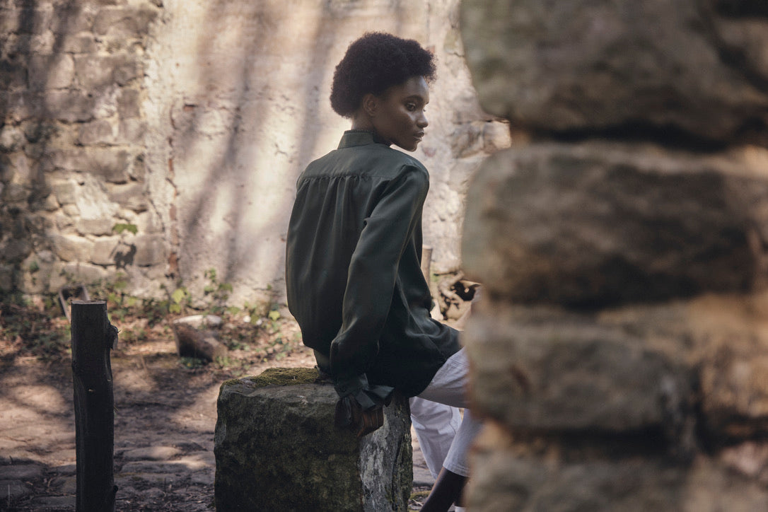 Chemise BOUDOIR Soie Kaki portée par une mannequin assise sur un bloc de pierre, de dos.