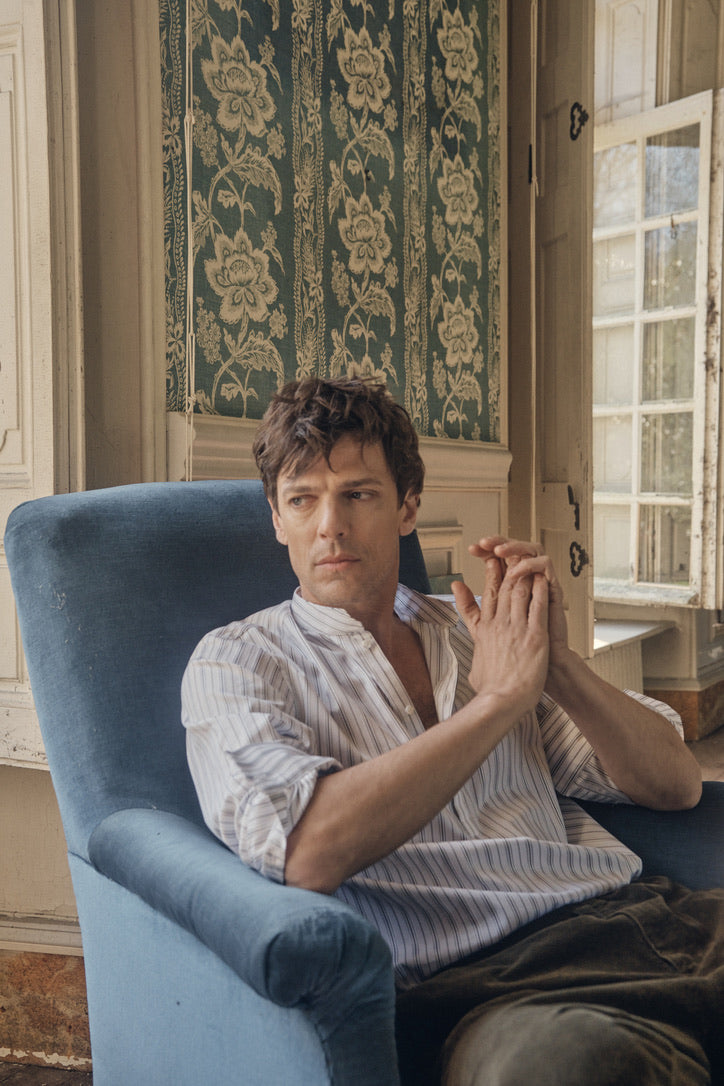Homme assis dans un fauteuil bleu à pampilles, le dos adossé à un mur orné de papier peint à motif floral, portant la chemise ÉPICURIEN Rayée Kaki en popeline de coton (coupe Bourrienne ample, col officier biseauté, poignets classiques retroussés), les mains jointes et le regard tourné vers la droite, dans une lumière douce.