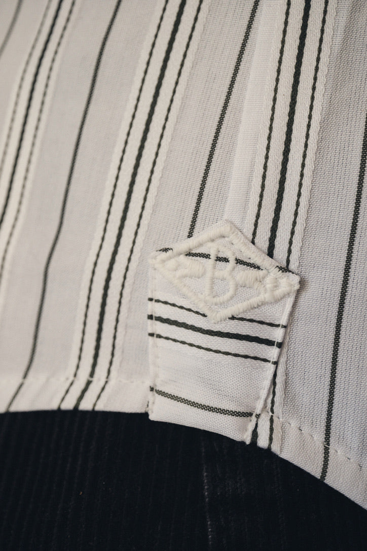 Gros plan sur l’ourlet latéral de la chemise, où le tissu en popeline kaki clair à fines rayures verticales blanches forme un petit pli net juste au-dessus du bord. Un empiècement en forme de losange déborde légèrement, reprenant le motif rayé et portant une broderie ton sur ton évoquant le monogramme Bourrienne Paris X. En arrière-plan, on devine un tissu sombre (pantalon en velours côtelé)