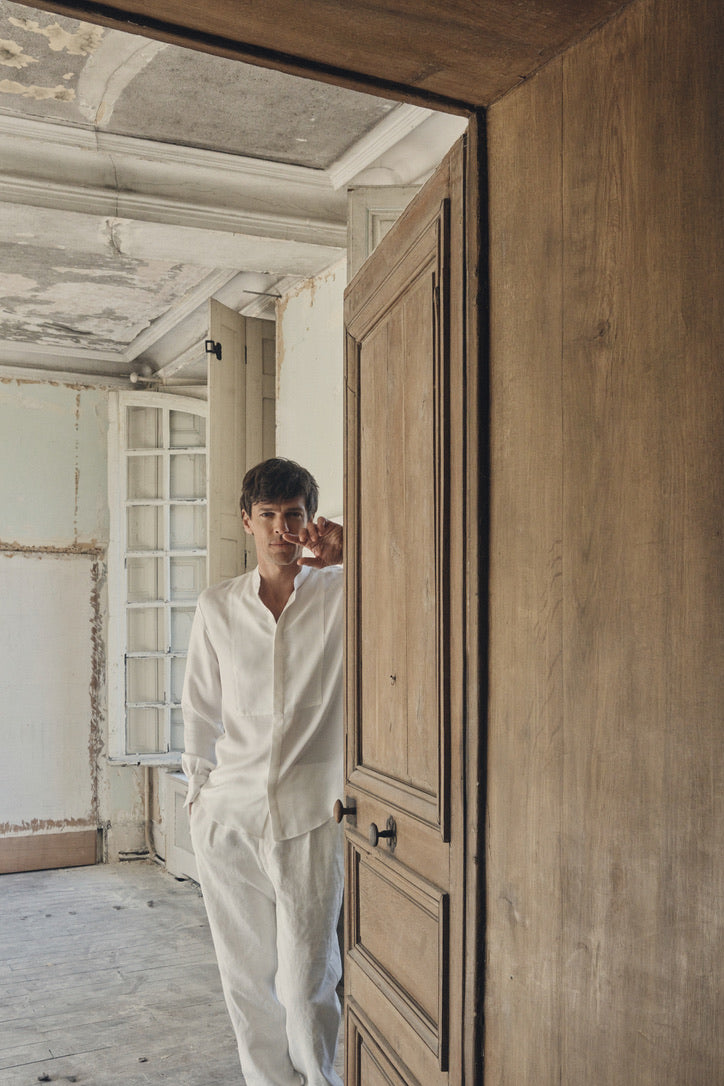 Homme de face, appuyé sur l’embrasure d’une porte en bois ancien, ajustant délicatement ses lunettes d’un geste nonchalant. Il porte la chemise NOCTURNE en soie blanche, col officier ouvert sur un plastron nid d’abeille et poignets Bourrienne signature, associée à un pantalon blanc cassé aux poches discrètes. Le décor patiné murs écaillés et parquet usé met en valeur la fluidité satinée de la soie.