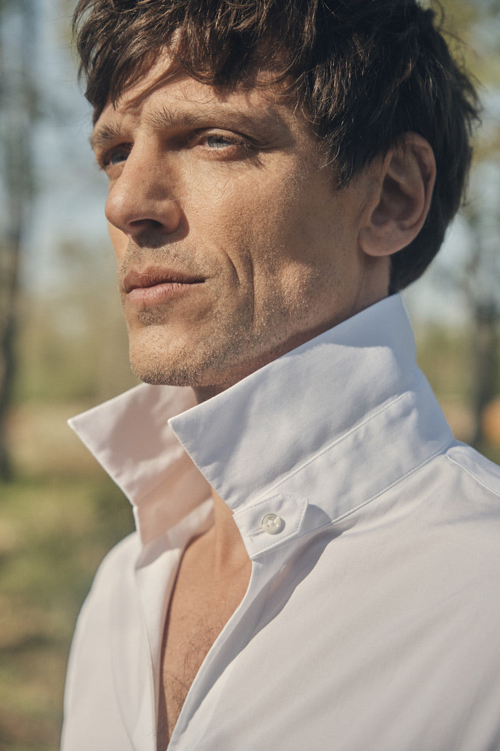 Photo plan poitrine d'un homme chatain, rasé, portant une chemise qui se nomme VOYAGE. Elle est en 100% Coton, fabriqué en Europe, une coupe Bourrienne, un col Français et des poignets Bourrienne. Sur la photo, le col de la chemise est levé