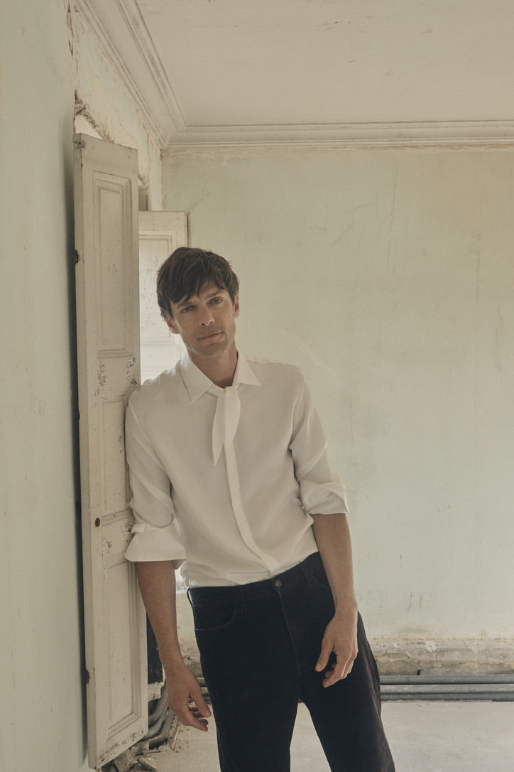 Homme appuyé contre une fenêtre à petits volets, portant la chemise PALETTE en soie blanche à coupe Bourrienne, col français et large nœud décontracté. Les manches retroussées dévoilent les poignets classiques, tandis que la soie satinée capte la lumière douce du jour. Le contraste entre le mur patiné et la fluidité du tissu souligne l’élégance moderne de la silhouette.