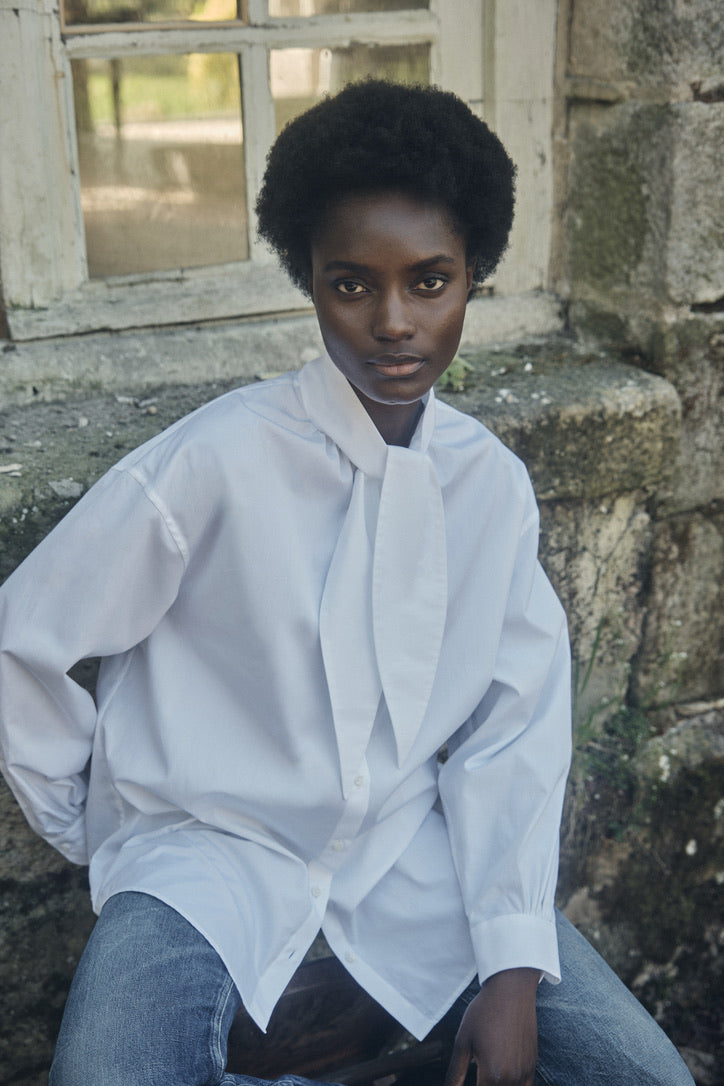 Femme assise portant la chemise MERVEILLEUSE, posant devant une maison en pierre.