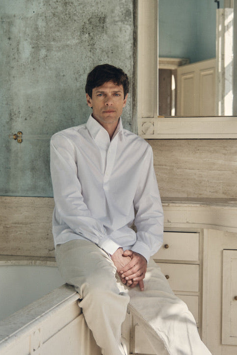 Une photo plan américain d'un homme assit sur le rebord d'une baignoire, il regarde l'objectif il porte une chemise 100% Coton, fabriqué en Europe, une coupe Bourrienne, un col Français et des Poignets Bourrienne