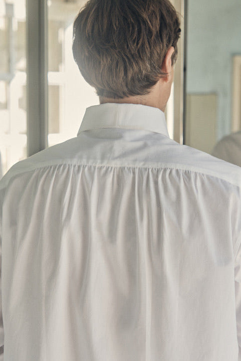 Une photo plan poitrine d'un homme de dos dans une salle de bain, il est face à un miroir, on voit son reflet. Il porte une chemise 100% Coton, fabriqué en Europe, une coupe Bourrienne, un col Français et des Poignets Bourrienne