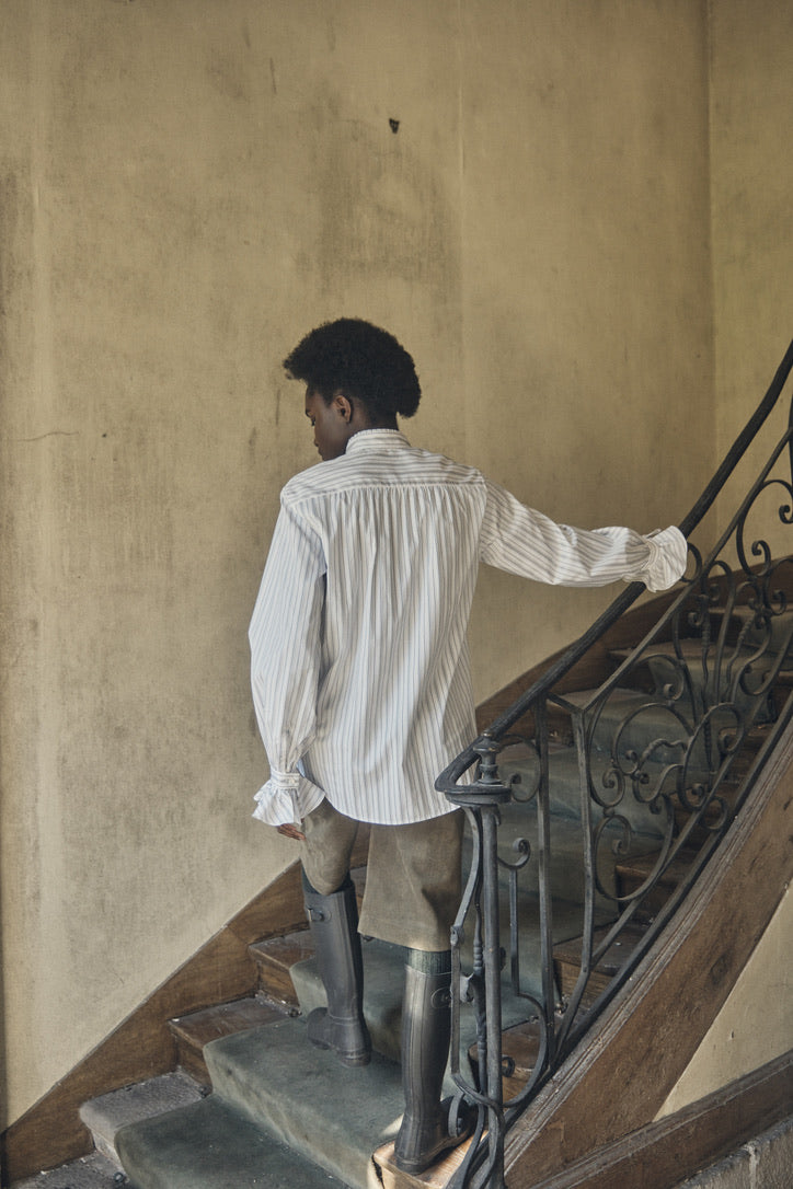 Femme posant de dos dans les escaliers d'une maison portant la chemise BOUDOIR Rayée Kaki, un bermuda kaki et des bottes hautes.