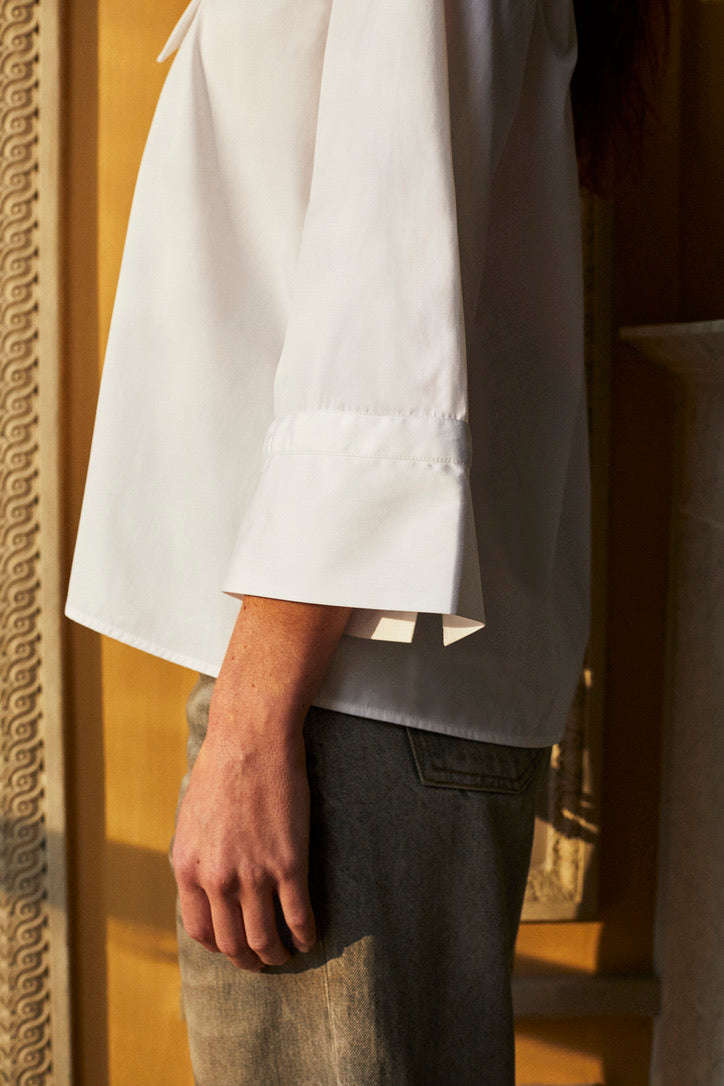 Photo verticale, plan taille, d'une femme de profil et debout. Elle porte un jean droit gris et une chemise blanche en popeline double retors italienne Bourrienne Paris X nommé GRACE. Une coupe avec un volume ajusté au niveau du buste et plus évasé en bas. Nous voyons très bien les manches ¾ avec des details brodés.