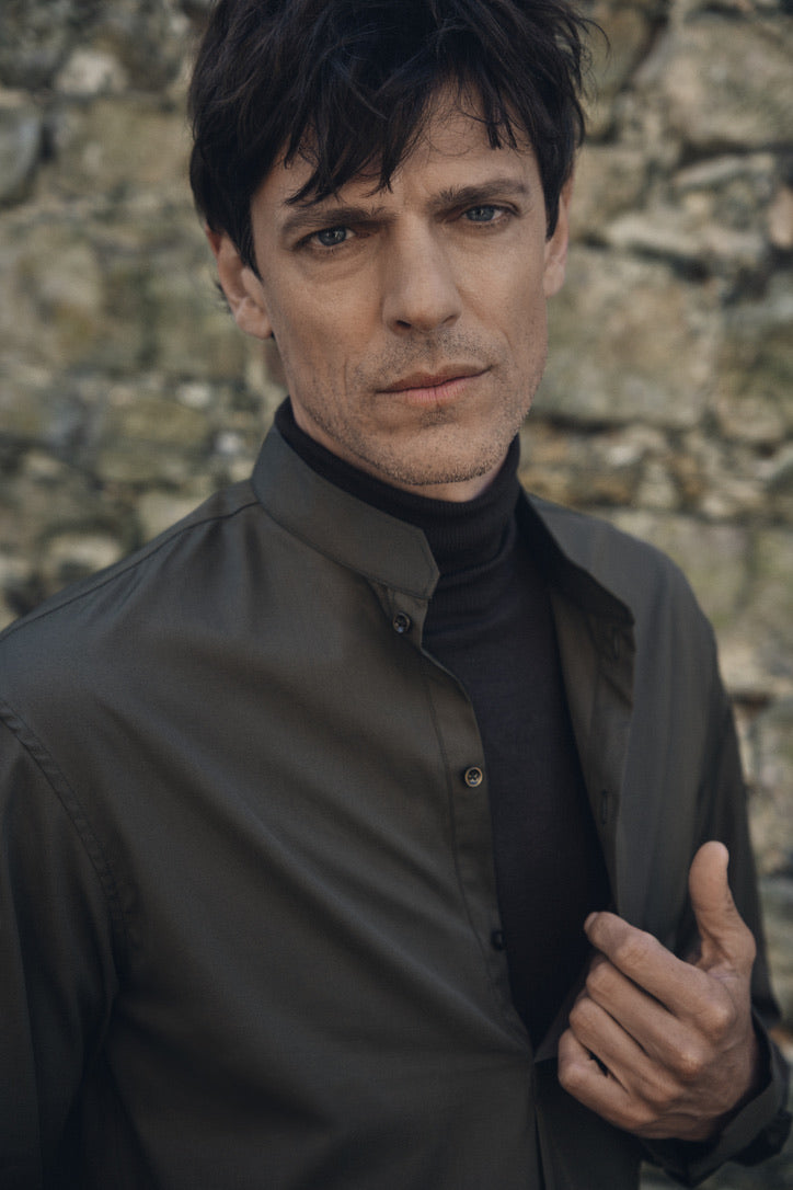 Gros plan sur un homme au regard pénétrant, ajustant le col officier biseauté de sa chemise ÉPICURIEN Kaki en popeline de coton. On distingue le liseré de la boutonnière et l’empiècement minimaliste, tandis que le fond flou de pierre évoque une ambiance rustique et élégante.