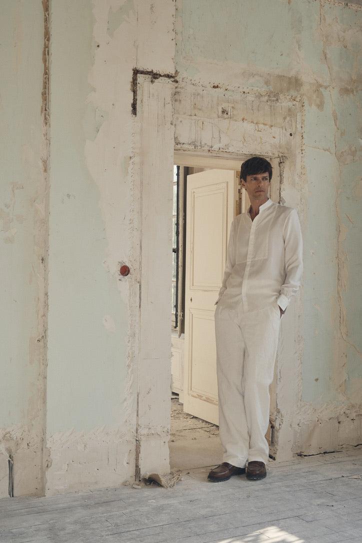 Homme appuyé dans l’embrasure d’une porte ancienne aux murs écaillés, mains en partie dans les poches d’un pantalon blanc cassé, portant la chemise NOCTURNE en soie blanche à col officier et plastron nid d’abeille. Les poignets Bourrienne, finement froncés et fermés par un bouton nacré, ressortent sous les manches légèrement retroussées. Le contraste entre la fluidité satinée de la soie et la texture brute du décor valorise la coupe ajustée et l’élégance poétique de la pièce.