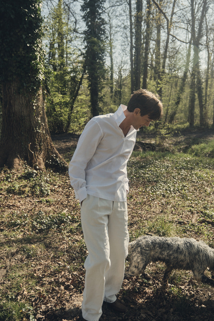 Photo pied pied d'un homme portant une chemise nommé VOYAGE, elle est en 100% Coton, fabriqué en Europe, une coupe Bourrienne, un col Français et des poignets Bourrienne. L'homme est debout dans la forêt, les mains dans ses poches, un chien marche à ses pieds.