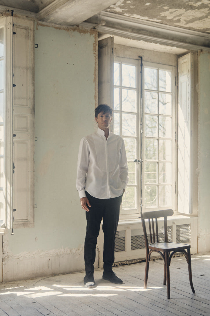 Photo plan américain d'un homme qui est debout, le visage face à la caméra, sa main gauche rentrée dans son pantalon, l'autre est relaché. Il porte une chemise blanche, coupe Bourrienne, poignets Français, col haut officier avec un plastron.