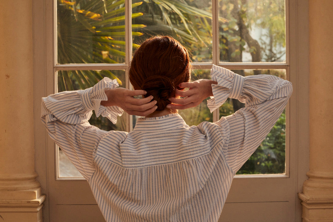 Photo horizontale d'une femme en chignon, rousse de dos. Elle porte une chemise Boudoir Seersucker Rayée Bleu Ses bras touche son chignon, nous apercevons plus en détails les volants des poignets, signature de la Maison Bourrienne. En face d'elle se trouve une fenêtre avec des palmiers.