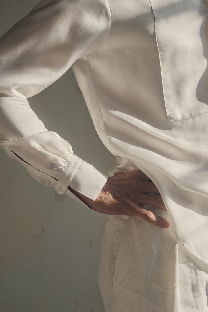 Gros plan sur le flanc droit d’un homme, main posée sur la hanche, révélant la coupe ajustée et le tombé fluide de la chemise NOCTURNE en soie blanche. On distingue le plastron en nid d’abeille parfaitement centré et le poignet Bourrienne froncé, fermé par un petit bouton nacré. La lumière rasante souligne la texture satinée du tissu et la finesse des surpiqûres.