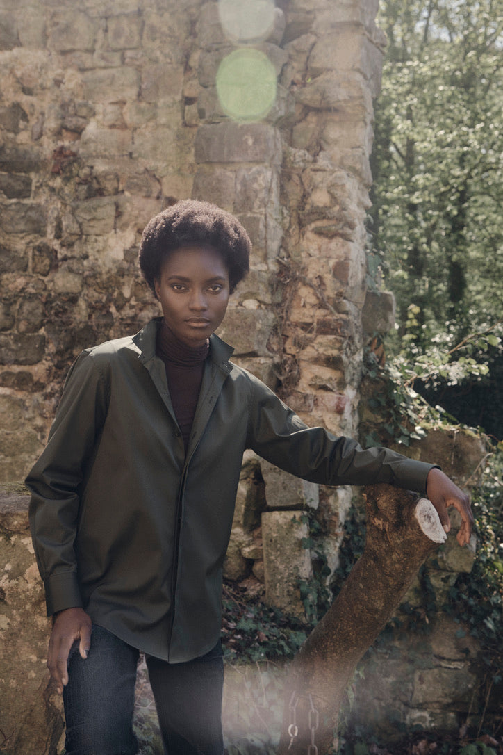 Plan américain d'une mannequin portant la chemise AMAZONE Kaki et un jean noir, dans un jardin devant un mur en pierres.