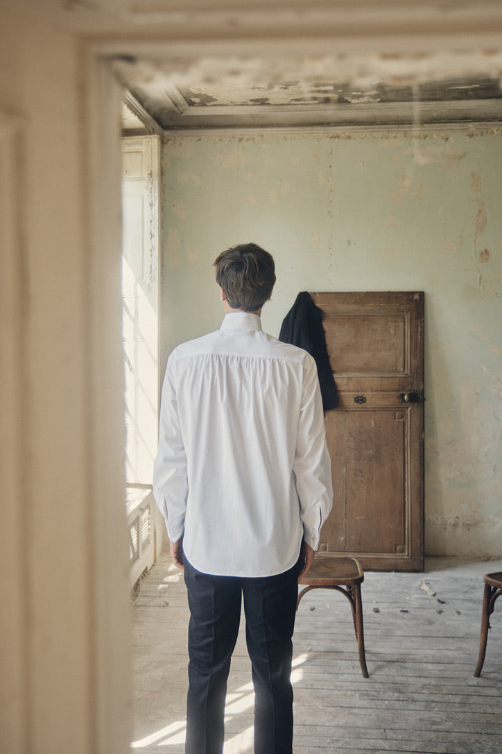 Photo plan américain d'un homme debout de dos. Il se trouve à l'intérieur d'un appartement, Il porte une chemise blanche, coupe Bourrienne, poignets Français, col haut officier avec un plastron.