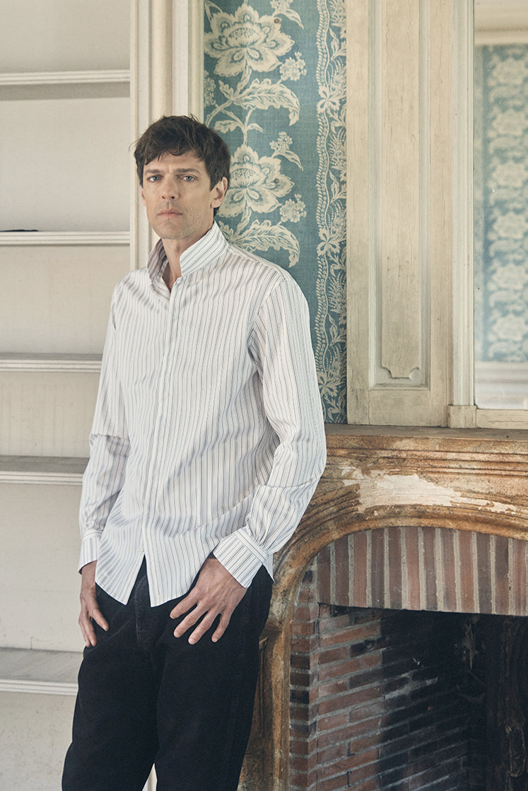 Homme de profil trois-quarts, regard tourné vers l’objectif, portant une chemise ajustée en popeline kaki à fines rayures verticales blanches, col montant fermé jusqu’au deuxième bouton, manches longues terminées par des poignets bouffants boutonnés. Il se tient debout, mains appuyées sur ses cuisses, devant une cheminée en pierre patinée et un mur recouvert d’un papier peint bleu à larges motifs floraux crème. Le décor ancien, aux moulures et boiseries usées, accentue l’élégance contemporaine de la tenue.