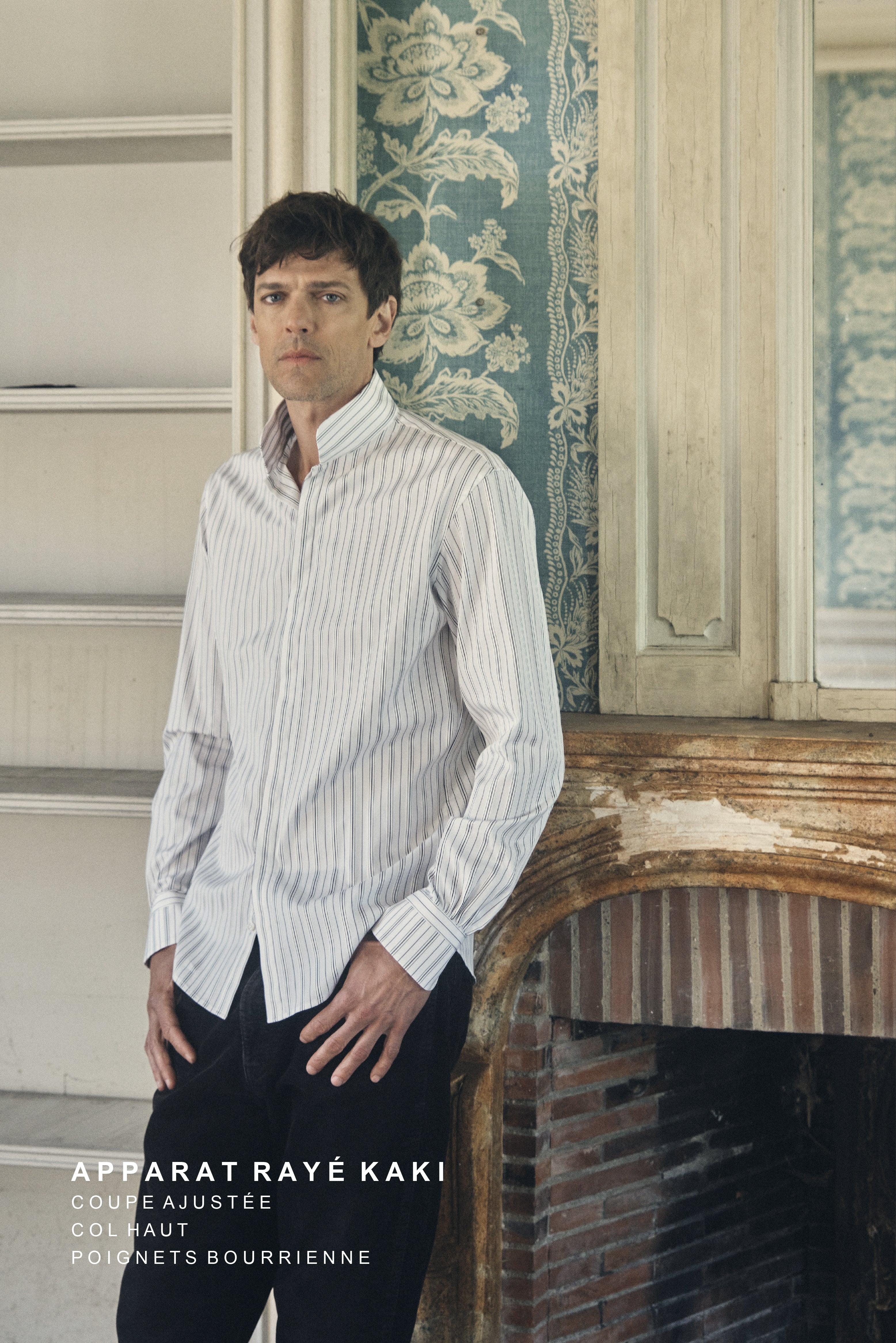Homme de profil trois-quarts, regard tourné vers l’objectif, portant une chemise ajustée en popeline kaki à fines rayures verticales blanches, col montant fermé jusqu’au deuxième bouton, manches longues terminées par des poignets bouffants boutonnés. Il se tient debout, mains appuyées sur ses cuisses, devant une cheminée en pierre patinée et un mur recouvert d’un papier peint bleu à larges motifs floraux crème. Le décor ancien, aux moulures et boiseries usées, accentue l’élégance contemporaine de la tenue.