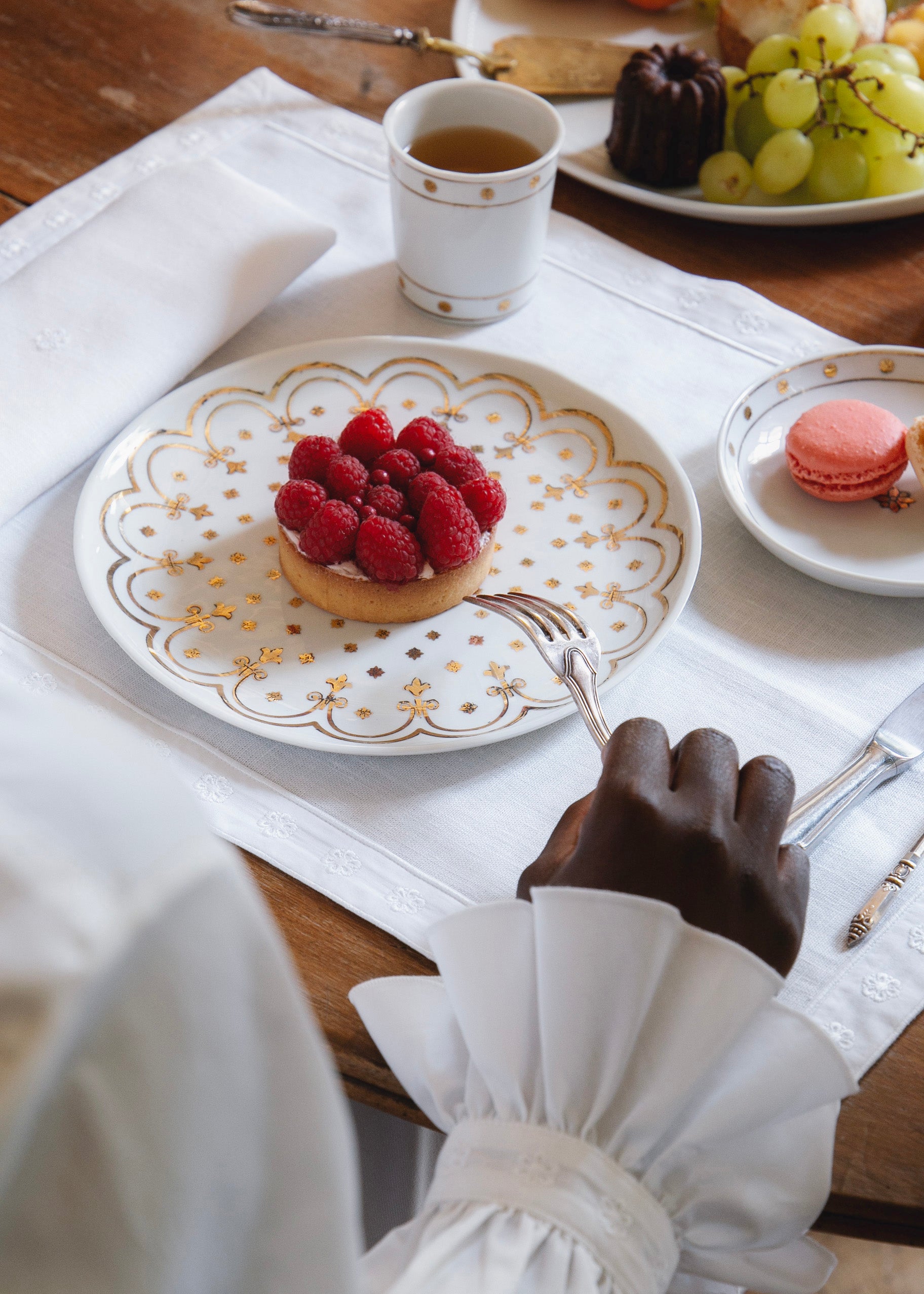 Une personne vêtue d’une chemise blanche à poignets volantés s’apprête à déguster une tartelette aux framboises, posée sur une assiette en porcelaine blanche ornée de motifs dorés. À côté, une tasse de thé, une coupelle avec un macaron rose et un plateau de pâtisseries et raisins complètent la scène sur une table dressée avec un set de table blanc.