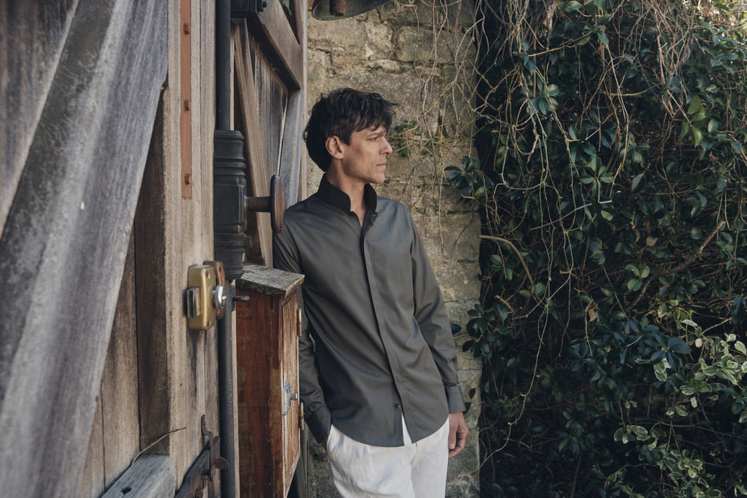 Un homme de profil trois-quarts, mains dans les poches de son pantalon blanc cassé à plis marqués, s’appuie contre une porte en bois patiné sous une cloche ancienne. Il porte une chemise ajustée kaki foncé en popeline, col montant ouvert et manches longues, dont la coupe cintrée met en valeur la fluidité du tissu. Le contraste entre la sobriété de la tenue et les textures brutes du mur de pierre et du lierre environnant crée une atmosphère à la fois rustique et élégante.
