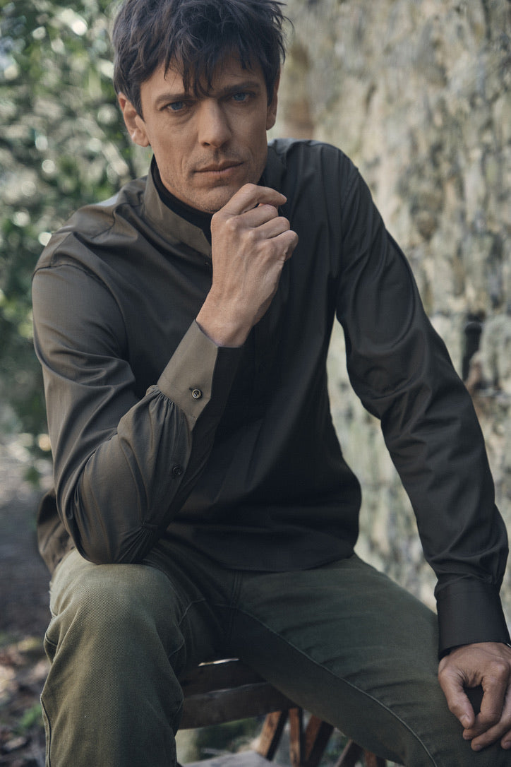 Homme assis sur un tabouret devant un mur en pierre, portant la chemise ÉPICURIEN Kaki en popeline de coton (coupe Bourrienne ample, col officier biseauté, poignets classiques) superposée à un col roulé noir, la main droite portée près du visage et le regard vers l’objectif.