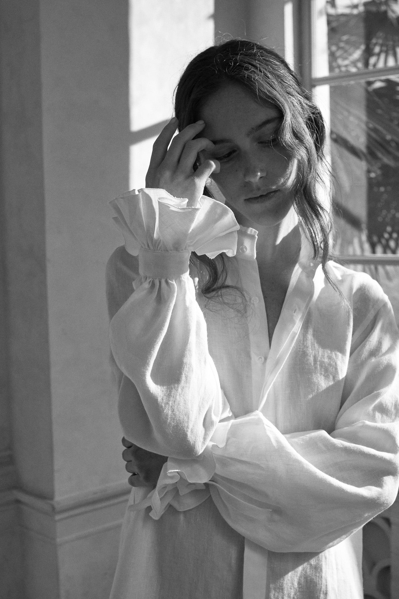 Photo en noir et blanc, plan américain d'une femme portant la chemise BOUDOIR dans un lin blanc plein de détails raffinés : un col haut subtilement bordé de petits volants pour une touche de sophistication et des manches légèrement bouffantes ornées de volants, qui tombent avec élégance sur la main. Sa main touche délicatement son visage, l'air pensive. La chemise confectionnée dans un lin belge.